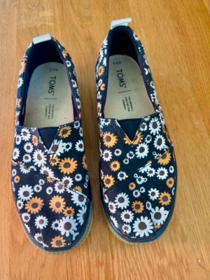 TOMS sneakers med blommigt mönster - Svarta sneakers från TOMS med vita och gula blommor över hela skon. Skorna har låg profil, elastisk detalj framtill och vit samt beige sula. Ovandelen är i textil och sulan är i gummi. Perfekta för dig som gillar färg och mönster.
