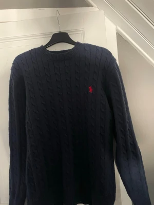 Mörkblå kabelstickad tröja Polo Ralph Lauren - Kabelstickad mörkblå tröja från Polo Ralph Lauren med rund halsringning och den klassiska röda logotypen broderad på bröstet. Tröjan har långa ärmar och ribbade muddar vid ärmslut och nederkant. Perfekt för lager på lager och stilren look.