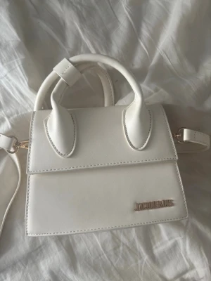 Vit Jacquemus handväska i läder - Snygg vit handväska från Jacquemus i stilren, boxig design med två handtag och axelrem. Väskan är gjord i slätt läder och har en metallogo framtill. Perfekt för dig som gillar trendiga accessoarer och minimalistisk stil.