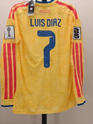 Colombia spelar Version långärmad matchtröja Adidas - Gul långärmad spelar Version fotbollströja från Adidas med blå detaljer och röda ränder på ärmarna. Officiell Colombia-tröja med nummer 7 och namnet Luis Diaz, samt FIFA World Cup-patchar på båda ärmarna. Broderad landslagslogga och modern passform.