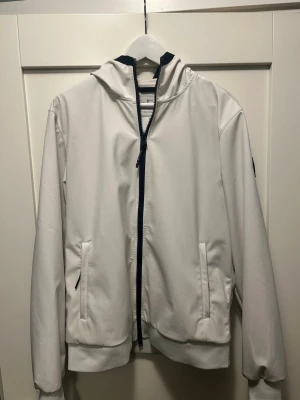 WOOLRICH Jacka - Snygg vit vindjacka från WOOLRICH med svart dragkedja och huva. Jackan har två fickor framtill och en svart logotyp på ärmen. Perfekt för dig som vill ha en stilren och sportig look. Materialet är lätt och vindtätt.