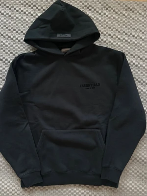 Essentials hoodie - Snygg svart hoodie från Essentials Fear of God med diskret logga på bröstet. Klassisk modell med huva och stor magficka. Perfekt för en avslappnad streetwear-look. Mjuk bomullskvalitet och ribbade muddar vid ärmslut och nederkant.