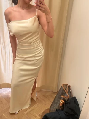 Balklänning  - Elegant långklänning i champagnefärgad satin med smala axelband och offshoulder-design. Klänningen har en snygg slits på sidan och figurnära passform som framhäver siluetten. Perfekt för dig som vill ha en stilren och trendig look. Använd 1 gång. Skickas 28 maj. strl:  S/36💕
