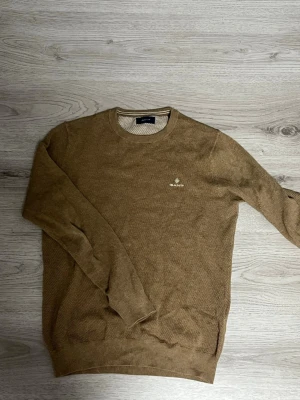 Beige stickad tröja från Gant - Snygg beige stickad tröja från Gant med rund halsringning och diskret logga broderad på bröstet. Tröjan har en klassisk passform och är tillverkad i ett mjukt, strukturerat material som passar perfekt till lager-på-lager-stil.