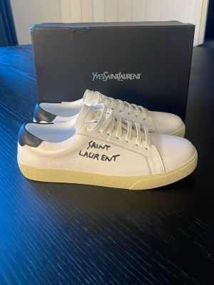 Vita Saint Laurent sneakers - Snygga vita sneakers från Saint Laurent med svart detalj på hälen och handskriven logga på sidan. Skorna har klassisk låg profil, vita snören och en beige sula. Tillverkade i läder för en exklusiv känsla och stilren look. Tveka inte på höra av er kan gå ner i pris på snabb affär 