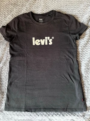 Svart Levi's t-shirt med logga - Klassisk svart t-shirt från Levi's med vit logga tryckt framtill. T-shirten har korta ärmar, rund halsringning och är gjord i mjuk bomull. En enkel och stilren tee som passar till allt och har den ikoniska röda Levi's-lappen i sidan.