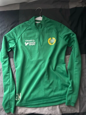 Grön träningströja Hammarby Craft - Grön långärmad träningströja från Craft med Hammarby IF-emblem på bröstet och Intersport-logga. Tröjan har halv dragkedja framtill och är tillverkad i ett mjukt, funktionsmaterial som passar perfekt för träning och sportaktiviteter.
