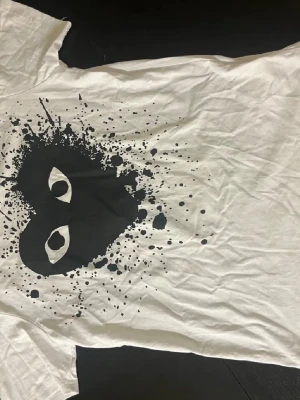 Vit t-shirt Comme des Garçons Play - Vit t-shirt från Comme des Garçons Play med svart grafiskt tryck framtill med färgstänk. Klassisk passform och rund halsringen och runt armarna. OBS!!!! Inte äkta!!!!