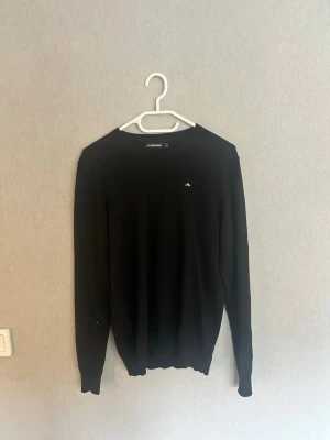 Svart stickad tröja från J.Lindeberg - Svart stickad tröja från J.Lindeberg med rund halsringning och diskret logga på bröstet. Tröjan har lång ärm och är tillverkad i ett mjukt material som passar perfekt till lager-på-lager-stil. Använd fåtal gånger, schyssta priser vid snabb affär.
