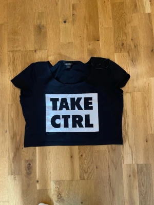 Svart croppad t-shirt Monki stl L bra skick - Svart croppad t-shirt från Monki med stort vitt tryck på magen där det står 'TAKE CTRL' i svarta bokstäver. T-shirten har rund halsringning och korta ärmar. Perfekt för en cool och avslappnad stil.
