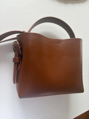 Brun väska från ARKET - Snygg brun bucket bag från ARKET i slätt läder. Väskan har ett stilrent och minimalistiskt uttryck med justerbar axelrem och handtag. Perfekt storlek för det viktigaste och med en cool knytdetalj på sidan. Enkel att matcha till olika outfits.