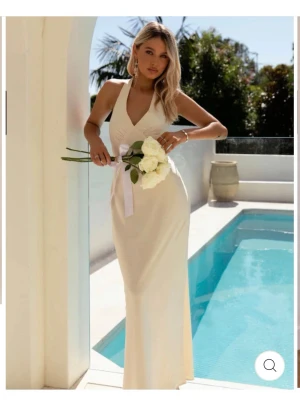 Champagnefärgad långklänning i satin - Elegant långklänning i champagnefärgad satin med djup v-ringning och öppen rygg. Klänningen har breda axelband som knyts i nacken och en smickrande figurnära passform. Perfekt för speciella tillfällen när du vill känna dig extra glam.