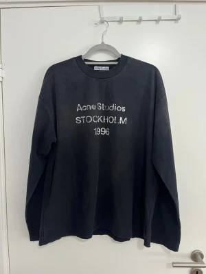 Svart långärmad Acne Studios S - Cool svart långärmad topp från Acne Studios med vit text 'Acne Studios STOCKHOLM 1996' framtill. Oversized passform och rund halsringning. Perfekt för en avslappnad och trendig look.