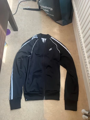 Svart Adidas hoodie med vita ränder - Svart hoodie från Adidas med dragkedja och klassiska vita ränder längs ärmarna. Tröjan har ribbade muddar och Adidas-logga på bröstet. Perfekt för en sportig och avslappnad stil.