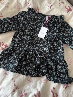Svart blommig volangblus med krage - Supersöt svart blus från Name It med småblommigt mönster i blått och vitt. Blusen har volangdetaljer, rundad krage och långa ärmar. Tillverkad i ett lätt och luftigt material som ger en skön känsla. Perfekt för dig som gillar romantisk och trendig stil.