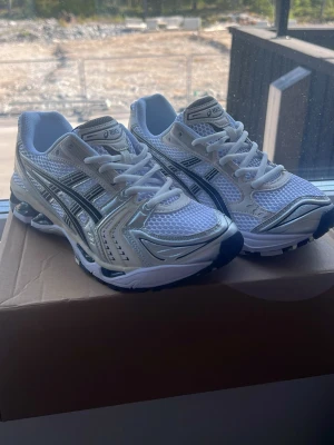 Vita ASICS Gel-Kayano 14 sneakers - Snygga vita ASICS Gel-Kayano 14 sneakers med silvriga detaljer och svarta ränder på sidorna. Ovandelen är i mesh och syntetmaterial för bra andningsförmåga och komfort. Sulan är vit med svarta och silvriga inslag, perfekt för en sportig och trendig look.