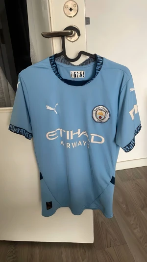 Manchester City matchtröja Puma blå - Snygg ljusblå Manchester City matchtröja från Puma med klubbmärke och Etihad Airways-tryck på bröstet. Tröjan har korta ärmar, rund hals med blått mönster och detaljer på ärmslut. Perfekt för fotbollsfans som vill visa sitt stöd.