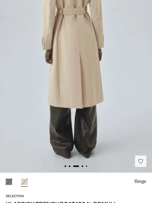 Beige klassisk trenchcoat bomull- oanvänd  - Stilren beige trenchcoat från Selection i 100% bomull. Dubbelknäppt med svarta knappar, bälte i midjan och klassisk krage. Långa ärmar med slejfdetalj vid ärmslut och två sidfickor. Ordinarie pris 1799kr 