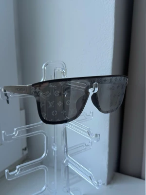  svarta solglasögon - Snygga svarta solglasögon  med monogrammönster på glasen. Modellen har en rak, futuristisk form och silvriga skalmar med ingraverad logotyp. Glasögonen har en modern och exklusiv look, perfekta för att sticka ut.