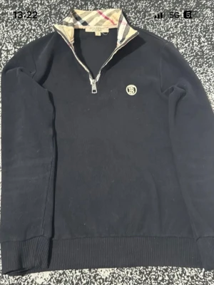 Svart Burberry tröja med dragkedja - Snygg svart tröja från Burberry med halv dragkedja och klassiskt rutigt mönster på insidan av kragen. Tröjan har ett diskret Burberry-emblem på bröstet och ribbade muddar vid ärmslut och nederkant. Perfekt för en stilren look.