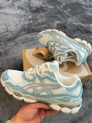 Asics GEL-NYC Blue - Fräscha sneakers från Asics i vitt och ljusblått med meshpaneler och detaljer i syntet. Klassisk låg modell med vita snören och silvriga detaljer på sidan. Perfekta för en sportig och trendig look.