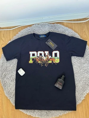 Svart Polo Ralph Lauren t-shirt - Snygg svart t-shirt med stort färgglatt POLO-tryck och polospelare på bröstet. Klassisk rund hals och korta ärmar. Perfekt för dig som vill ha en ikonisk och sportig look.