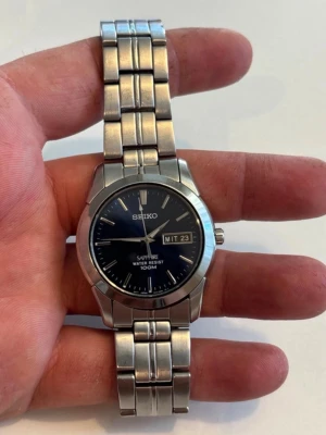 Seiko silverfärgad klocka med länkarmband - Stilren Seiko klocka i silverfärgat rostfritt stål med svart urtavla och dag- och datumvisning. Klockan har ett klassiskt länkarmband och tydliga indexmarkeringar. Vattentät upp till 100 meter och har safirglas för extra hållbarhet.