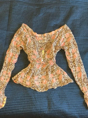 Blommig volangblus med knappar - Supersöt blus med blommigt mönster i rosa och brunt. Flowy modell med volanger och dekorativa knappar framtill och vid ärmslut. Långa ärmar och romantisk känsla, perfekt för dig som gillar vintage vibes. Tunn och lätt i materialet.