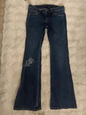 Blå bootcut jeans med slitning - Snygga blå bootcut jeans med klassisk femficksdesign och slitning på vänster ben. Jeansen har normal midja och är tillverkade i denim med lätt stretch. Perfekta för en avslappnad och trendig look.