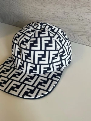 Svartvit Fendi keps med FF-mönster - Säljer en riktigt snygg svartvit keps från Fendi med det klassiska FF-logomönstret över hela kepsen. Kepsen har rak skärm och är tillverkad i ett slitstarkt tyg som känns lyxigt. Perfekt för dig som vill sticka ut med en ikonisk accessoar.