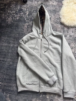 Grå hoodie från Burberry med dragkedja - Snygg grå hoodie från Burberry med dragkedja och klassiskt rutigt foder i huvan. Hoodien har fickor framtill, ribbade muddar och justerbar dragsko i huvan. Perfekt för en chill och stilren look.