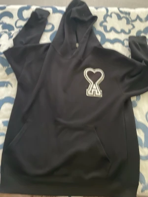 Svart hoodie med hjärtdetalj - Säljer en svart hoodie med stor huva och magficka. På bröstet finns en vit broderad detalj i form av ett hjärta och bokstaven A. Hoodien har en avslappnad passform och är perfekt för en chill look. Materialet känns mjukt och bekvämt.