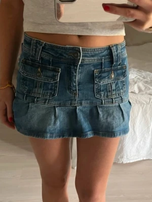 Blå jeanskjol med fickor - Snygg blå jeanskjol i kort modell med två stora fickor framtill och markerad söm nedtill. Kjolen har bälteshällor och stängs med knapp och dragkedja. Perfekt för en trendig och avslappnad look.