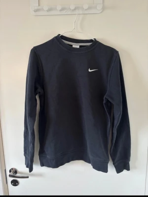 Svart sweatshirt från Nike - Klassisk svart sweatshirt från Nike med rund halsringning och vit broderad logga på bröstet. Tröjan har långa ärmar och ribbade muddar vid ärmslut och nederkant. Perfekt för en avslappnad och sportig stil.