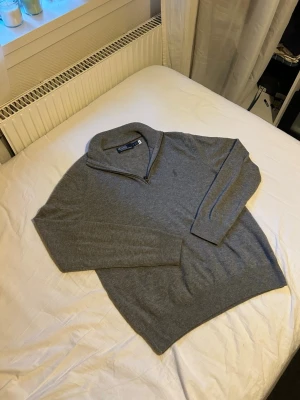 Grå halvzip-tröja från Polo Ralph Lauren - Klassisk grå stickad tröja från Polo Ralph Lauren med halv dragkedja och broderad logga på bröstet. Tröjan har lång ärm, ribbade muddar och är tillverkad i mjukt material som passar perfekt till lager-på-lager-stil. Självklart äkta!