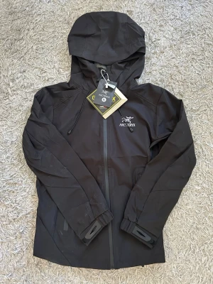 Svart regnjacka  - Snygg svart regnjacka med huva och dragkedja framtill. Jackan är tillverkad i slitstarkt Gore-Tex-material som står emot regn och vind. Justerbara muddar och minimalistisk design med logga på bröstet. Perfekt för blöta dagar. UTAN LAPPAR.