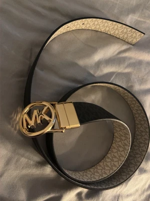 Svart Michael Kors bälte med guldspänne - Snyggt svart/vitt bälte från Michael Kors med ett stort, runt guldspänne med MK-logga. Man kan skifta mellan den svarta och den vita färgen. Helt ny och oanvänd. Ungefär 85 cm i längden.