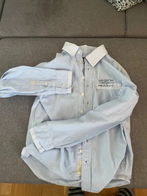 Blårandig skjorta från H&M - Snygg blårandig skjorta från H&M med vit krage samt text på bröstfickan. Ej trasig, är lite gammal men funkar utmärkt eftersom jag inte har använt den många gånger. Perfekt till jeans eller chinos för en clean look. Passar perfekt till sommar.