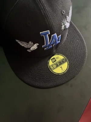 New Era keps - Snygg svart keps från New Era 59FIFTY med LA-logga i blått framtill, broderade vita fåglar och MLB-logga bak. Klassisk flat brim och stilren design, perfekt för dig som gillar streetwear och sportiga accessoarer.