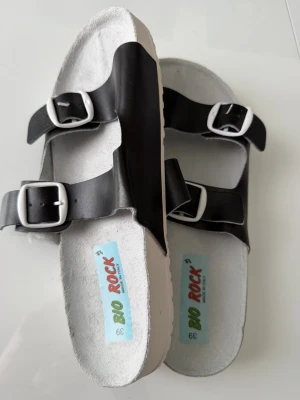 Svarta sandaler från Bio Rock - Stilrena svarta sandaler från Bio Rock med två justerbara remmar och silvriga spännen. Sulan är vit och har ett vågigt mönster undertill för extra grepp. Ovandelen är i syntetmaterial och innersulan är ljus för en fräsch look.
