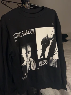 Svart Tupac Shakur sweatshirt - Cool svart sweatshirt med tryck av Tupac Shakur i vitt. Tröjan har rund halsringning och långa ärmar. Stort grafiskt print framtill med text och bilder på Tupac. Perfekt för dig som gillar streetwear och hiphop.