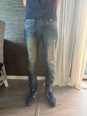 Blå slitna jeans från Scotch & Soda - Säljer ett par blå jeans från Scotch & Soda med slitningar och tvättade detaljer. Jeansen har smal passform, klassisk femficksmodell och en röd detalj vid fickan. Perfekta för en avslappnad och trendig look.