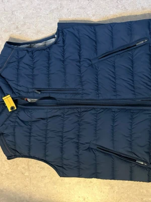 Mörkblå dunväst från Parajumpers - Snygg mörkblå dunväst från Parajumpers med gul detalj vid kragen och flera dragkedjeförsedda fickor. Ända lilla lilla slitaget på den är lappen på  dragkedjan, limmar man så är den som ny 