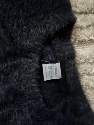 Mörkblå fluffig stickad tröja Lindex - Mysig mörkblå stickad tröja från Lindex med fluffig textur och broderad text 'Merci Paris' framtill. Tröjan har rund halsringning och långa ärmar, perfekt för att hålla värmen under kyliga dagar.