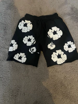 Denim Tears Shorts | Svart - Svarta shorts från Denim Tears med vita blommiga mönster och snörning i midjan. I storlek M, passar mig som 185 cm bra. De har en avslappnad passform och är perfekta för varma dagar. Materialet känns mjukt och bekvämt, och designen sticker ut med sina stora blommor och perfekta inför denna sommaren. Pris kan diskuteras. Knappt använd