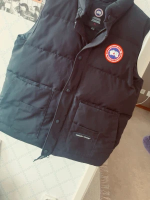Svart dunväst från Canada Goose. - Hjälper min vän att sälja hans iprincip helt nya väst, använd typ 4 gånger men legat i en garderob i ca 1 år. Finns inga skavanker på västen.