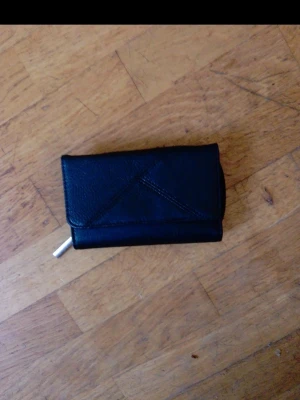 Svart plånbok i läderlook - Snygg svart plånbok med stilren design och triangulär detalj på framsidan. Plånboken har en praktisk dragkedja och är tillverkad i material som liknar läder. Perfekt för att hålla koll på kort och kontanter.