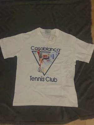Vit Casablanca Tennis Club t-shirt - Vit t-shirt från Casablanca med trycket 'Tennis Club' och grafiskt motiv framtill. Klassisk rund hals och korta ärmar. Tillverkad i mjuk bomull och har en clean, sportig vibe som passar perfekt till streetwear eller casual outfits.