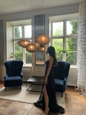 Svart långklänning med slits - Säljer en elegant svart långklänning med strapless design och hög slits på sidan. Klänningen har 3D-blommiga detaljer över hela tyget som ger en lyxig känsla. Perfekt för bal eller festliga tillfällen. Materialet känns som polyester.