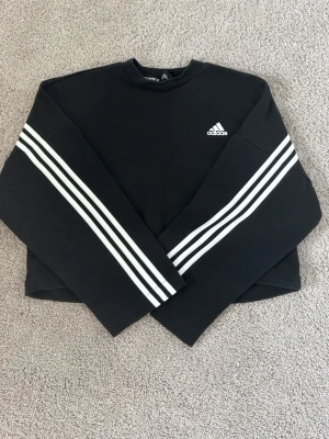 Svart sweatshirt från Adidas - Säljer en svart sweatshirt från Adidas med klassiska vita ränder längs ärmarna och Adidas-logga på bröstet. 